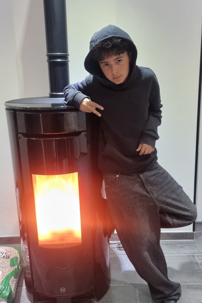Garçon en hoodie noir UN1K appuyé contre un poêle à bois allumé, posant avec un style décontracté et urbain.